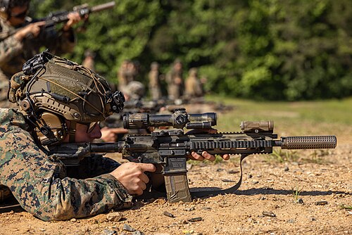 A Marine in modern camouflage and tactical gear in the prone position on a firing range, aiming a rifle equipped with a large scope and suppressor.