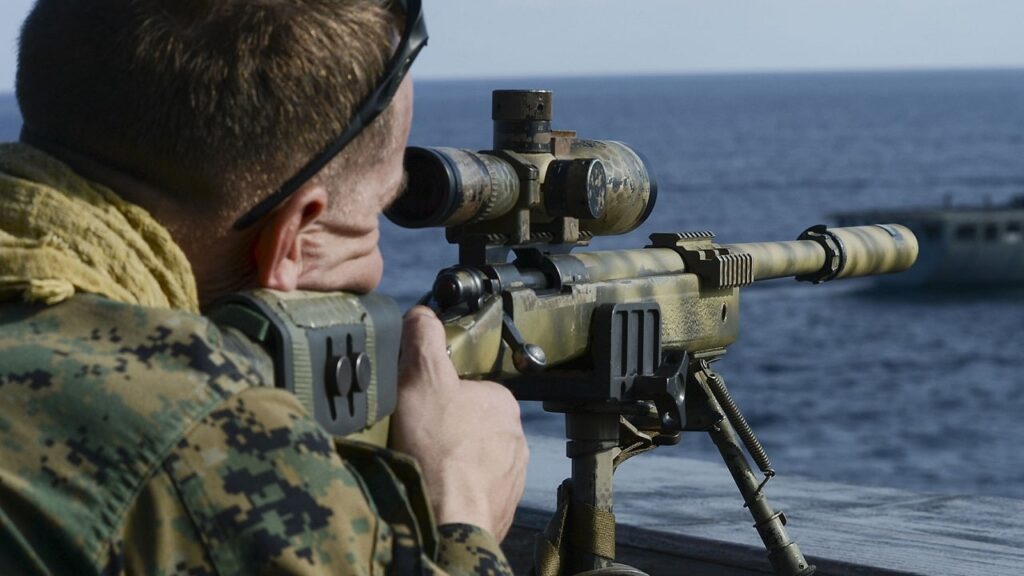 Marine in a camouflage uniform is seen from behind, aiming a large sniper rifle with a scope and bipod over the water, with a ship visible in the background