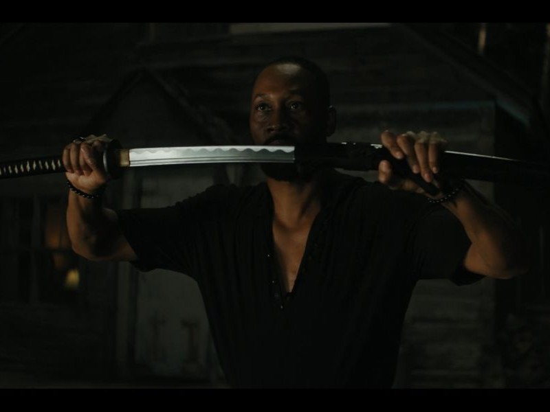A person dressed in black holding up a sword with both hands, the sword halfway out of the sheath.