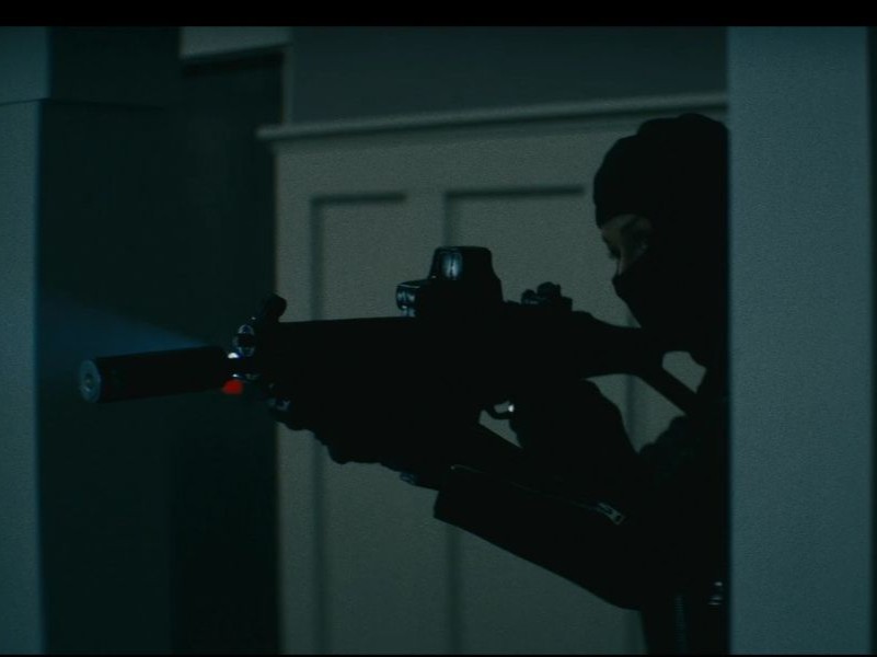 A person dressed in all black, pointing a rifle with a silencer on it.