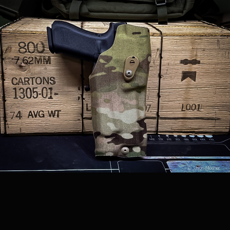 A black handgun in a camo holster, leaning up against a wooden box.
