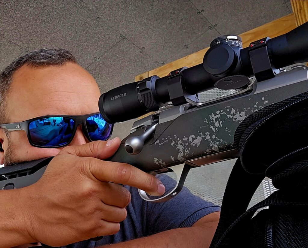 A man wearing sunglasses takes aim with a hunting rifle at a shooting range