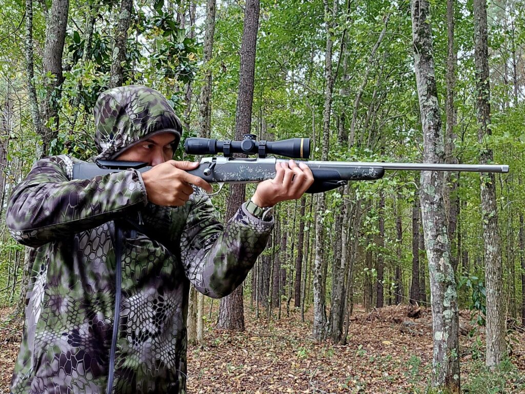 A man in camouflage holding a rifle in a standing shooting position in the forest