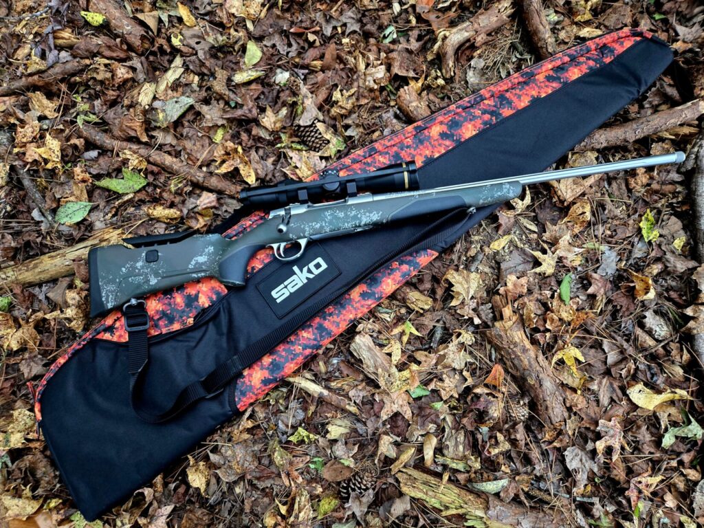 A hunting rifle with scope and scope caps on top of an orange camouflage case in a woodland backdrop