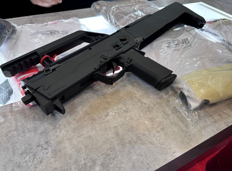 A black gun laying on a table with a long handle that runs across the top so it can fold into a "box".