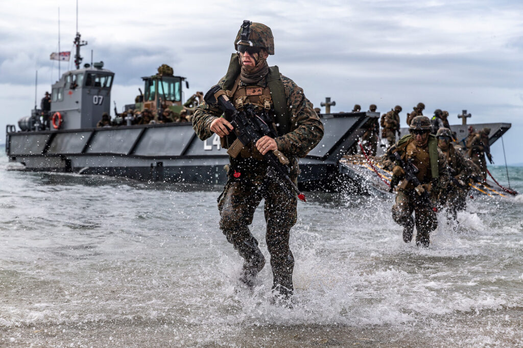 A group of Marines in combat gear are rushing out of the ocean and onto a beach during an amphibious landing exercise, with a landing craft utility (LCU) boat in the background.