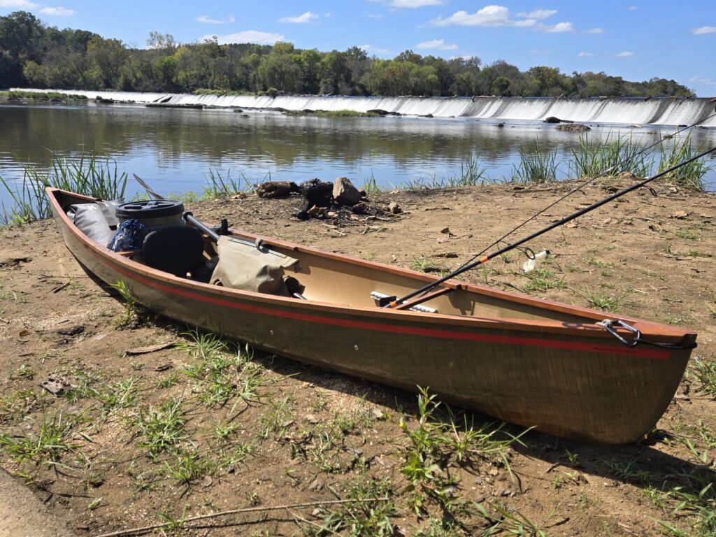 An ultralight canoe on a river bank with dry bags, fishing poles, and watertight bucket