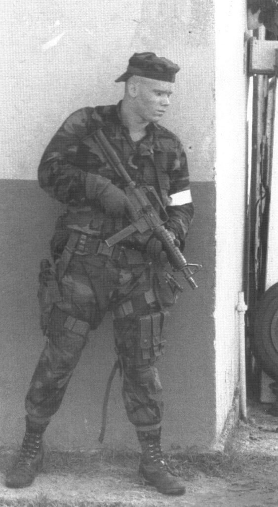 Black and white full-body shot of a Marine in woodland camouflage and a cap, armed with Colt SMG standing against a corner of a building.