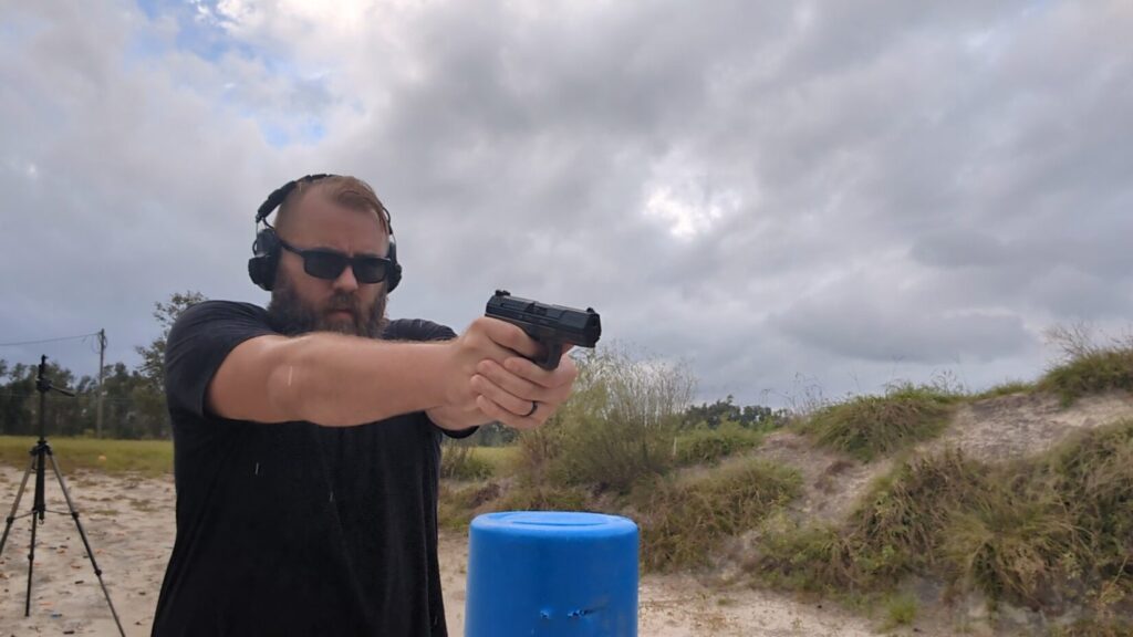 Close-up of a man with a beard and sunglasses aiming a pistol with an attached weapon light.