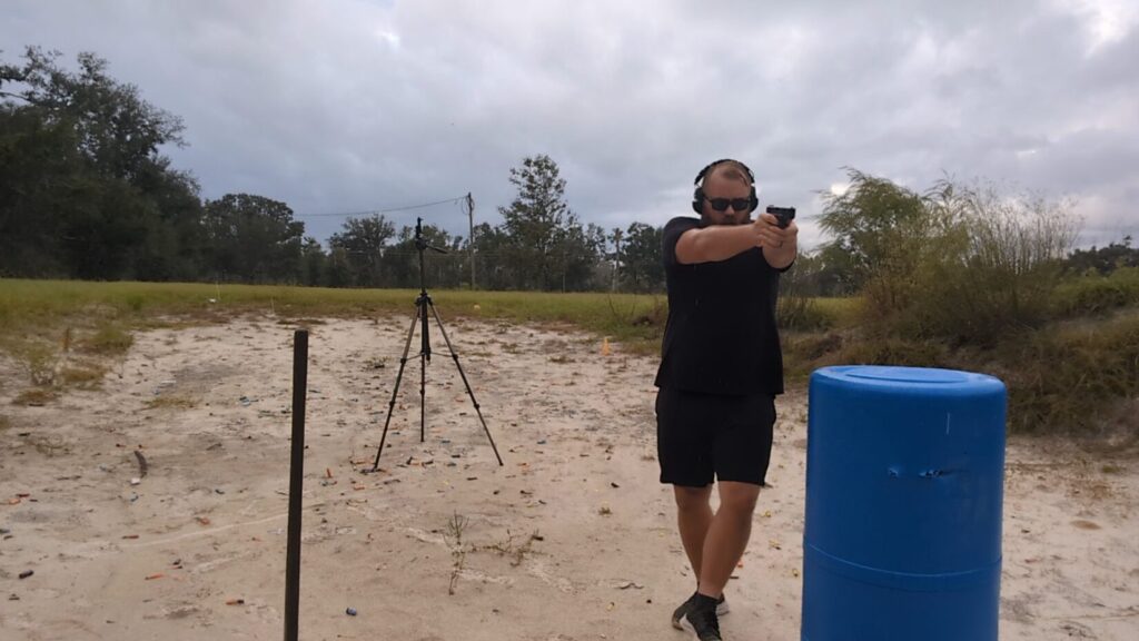 Man stepping around a blue barrel while aiming a pistol