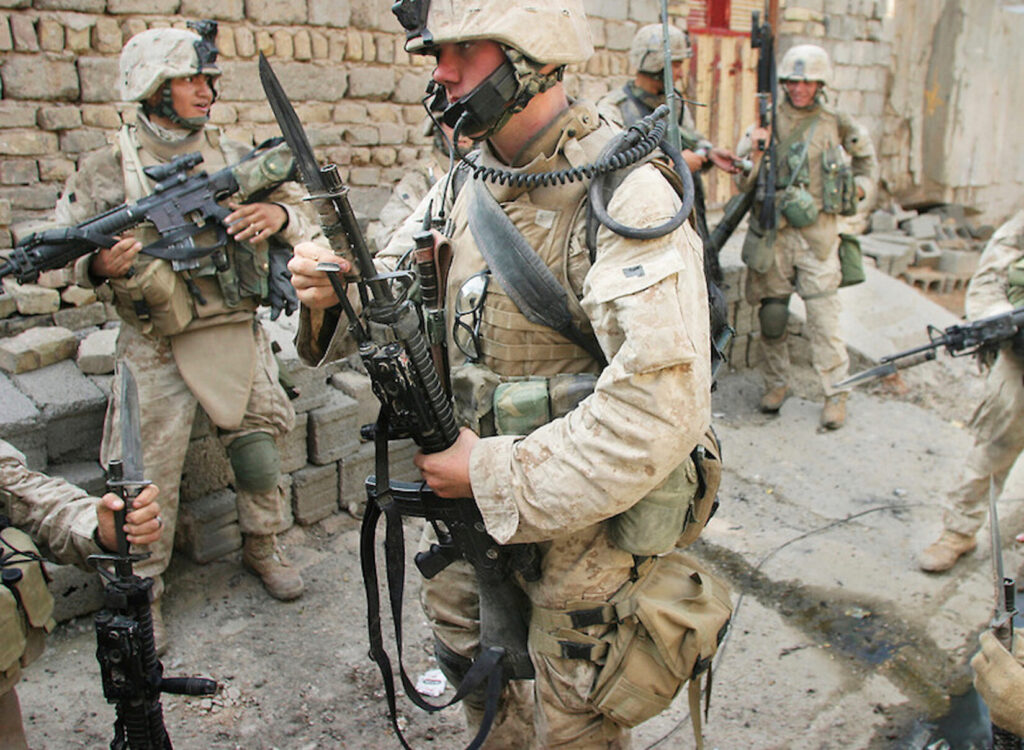 Marines in desert camouflage and combat gear stand in a rubble-filled urban environment. The Marine in the center is holding an M16 with a bayonet fixed.