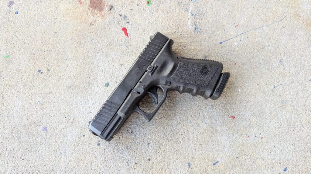 A Glock 19 black polymer handgun with an extended magazine base plate rests diagonally on a concrete floor speckled with paint.