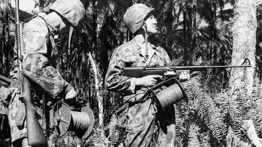Black and white image of two Marines in a jungle environment, wearing camouflage fatigues and helmets. One holds a Johnson light machine gun.