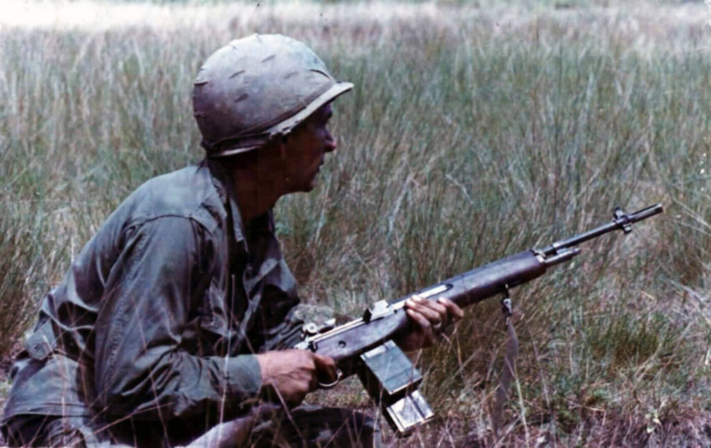 American soldier in Vietnam kneeling in tall grass, holding an M14 rifle.