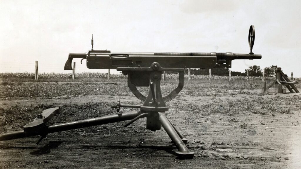 black and white image of a large, long-barreled machine gun mounted on a tripod, resting on the ground.