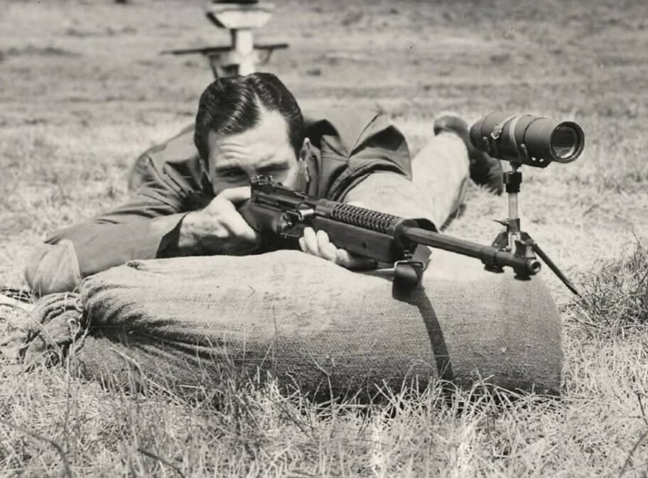 Black and white photo of a man in a prone position aiming a rifle over a sandbag, with a spotting scope mounted on a tripod to his right.