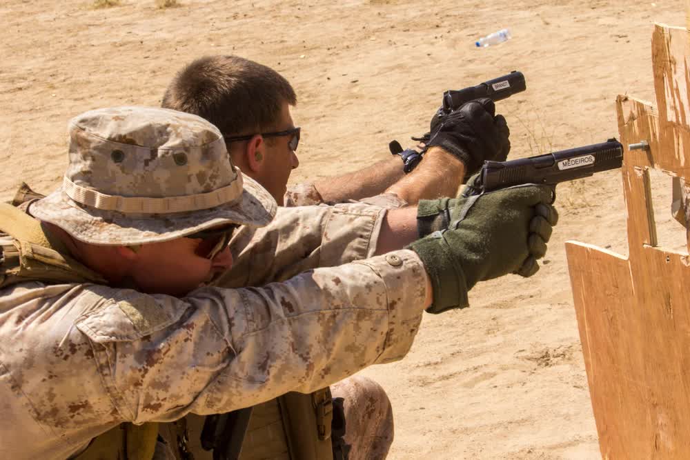 Two individuals in desert camouflage and boonie hats are on a firing range, both aiming pistols at a target.