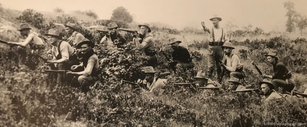 Black and white historical photo of a group of Marines aiming rifles from a prone position in a brushy, hilly area, with a man standing and giving a command.