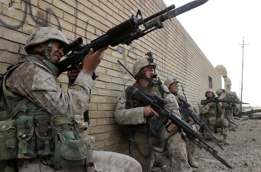 A group of Marines in desert camouflage taking cover behind a brick wall during a patrol. The Marine in the foreground is aiming an M16 rifle with a bayonet attached.