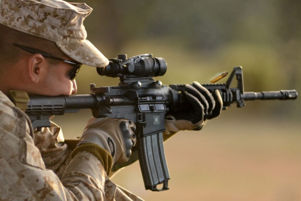 A close-up of a Marine in uniform and sunglasses firing an M4 carbine with an optical sight, with a spent casing ejecting from the weapon.