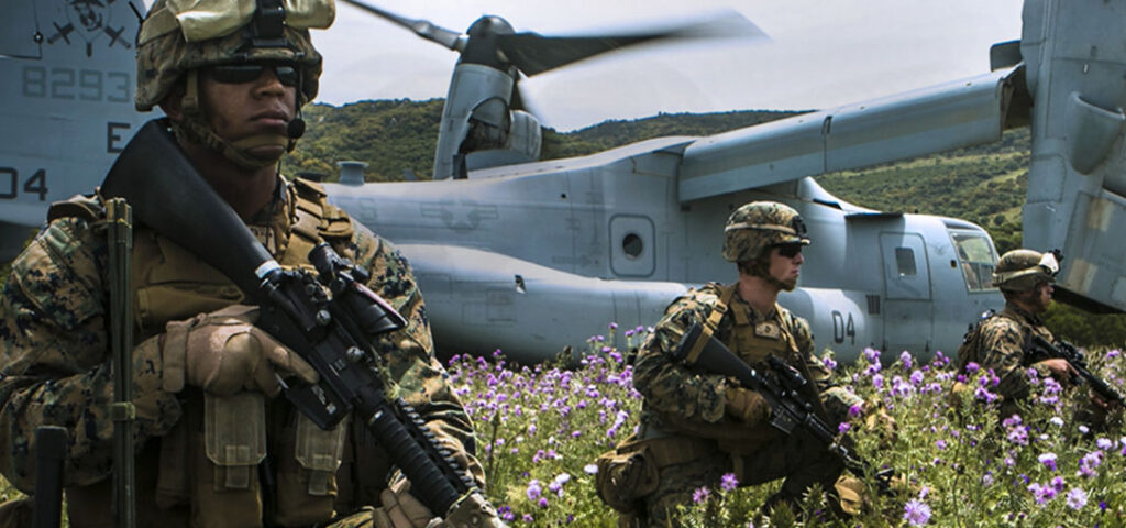Three Marines in tactical gear and sunglasses are positioned among purple wildflowers in a field, with a V-22 Osprey tiltrotor aircraft visible behind them on a hillside.