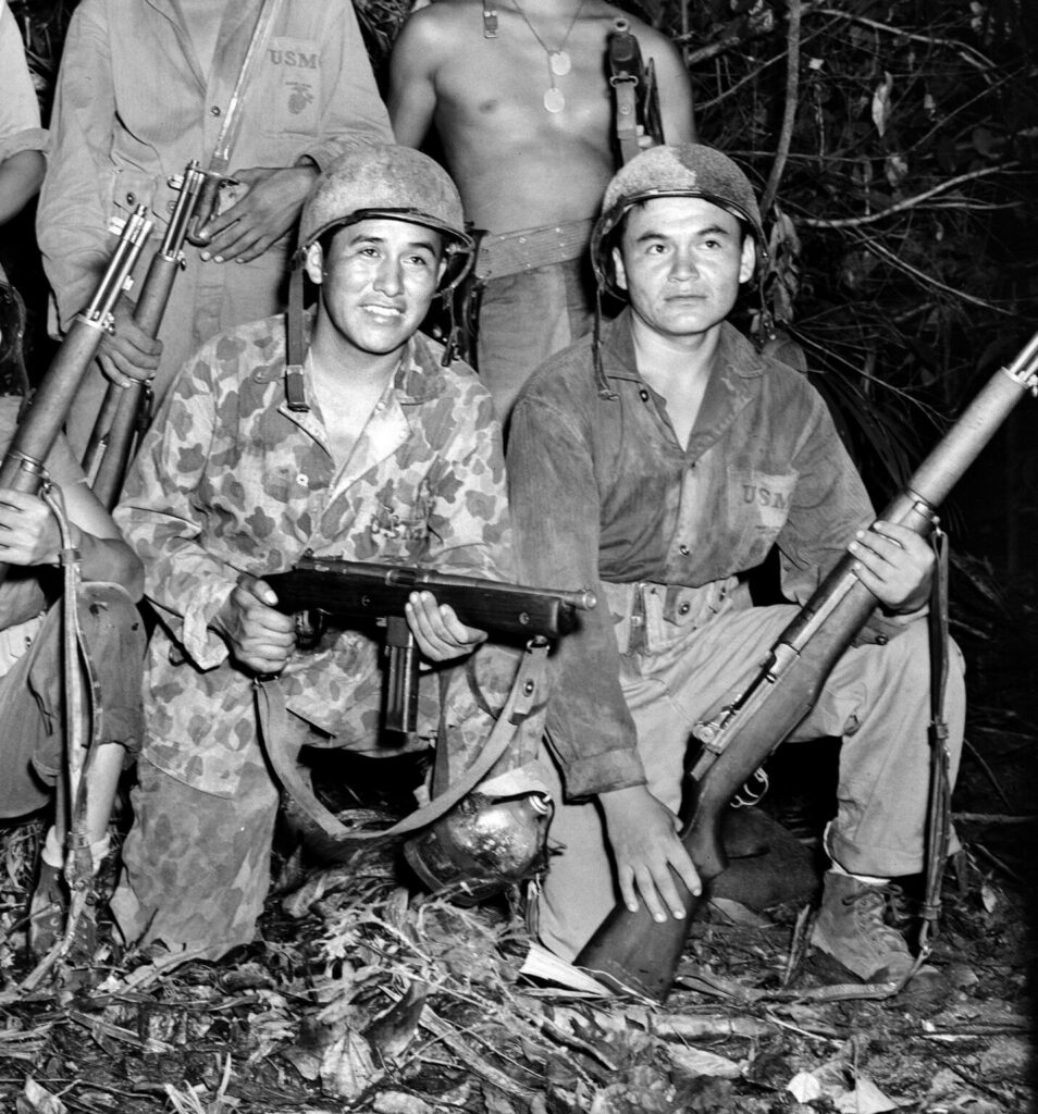 Black and white photo of two Marines smiling while kneeling in a jungle setting. One holds a submachine gun and the other holds a rifle.