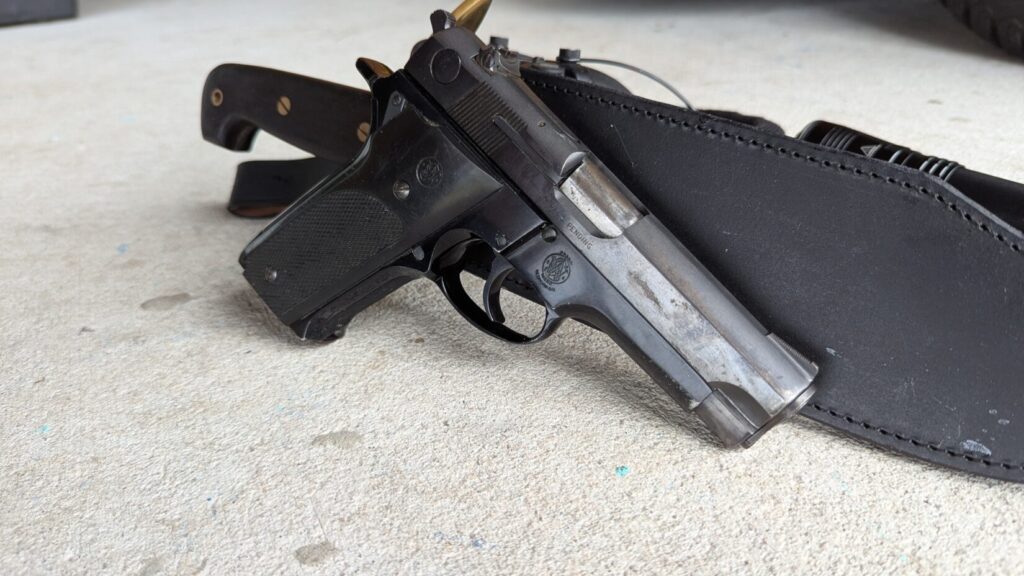 Close-up of the Smith & Wesson Model 59 pistol with a heavily worn slide, resting against a black leather knife sheath and a large bowie knife handle.