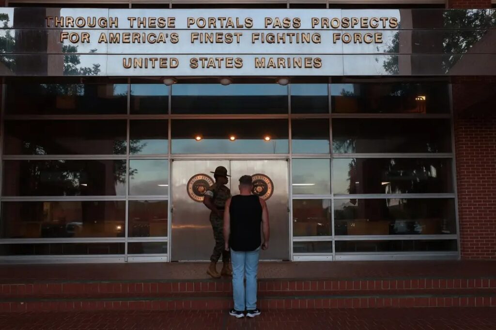 A potential recruit stands facing the glass entrance doors of a Marine Corps Recruit Depot receiving building, with the inscription "THROUGH THESE PORTALS PASS PROSPECTS FOR AMERICA'S FINEST FIGHTING FORCE UNITED STATES MARINES" above the doors.
