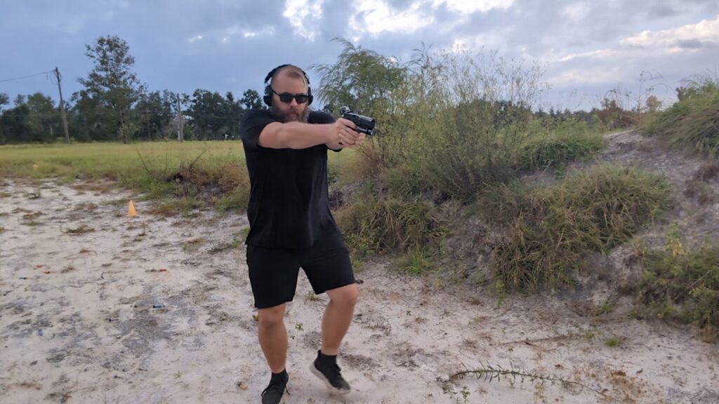 Man aiming a pistol in an outdoor range, with one foot slightly lifted.