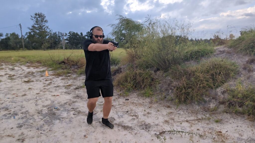 Man in shooting gear aiming a pistol while taking a step forward.