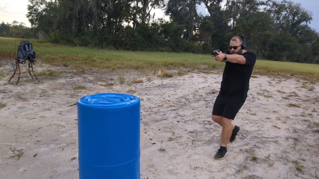 Man aiming a black pistol at an outdoor range, standing in front of a blue barrel.