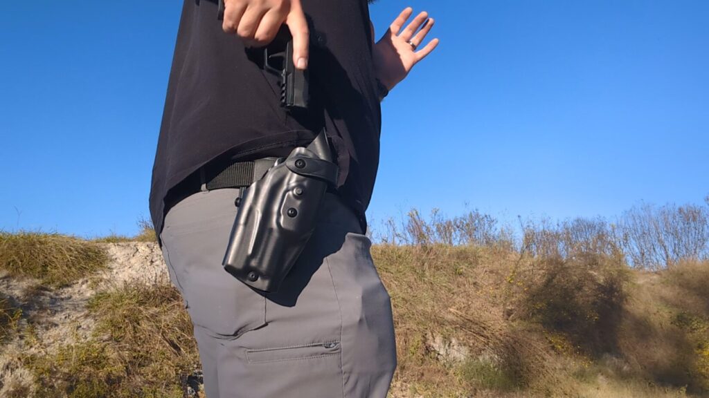 Man re-holstering a handgun into a black, hip-mounted retention holster outdoors.