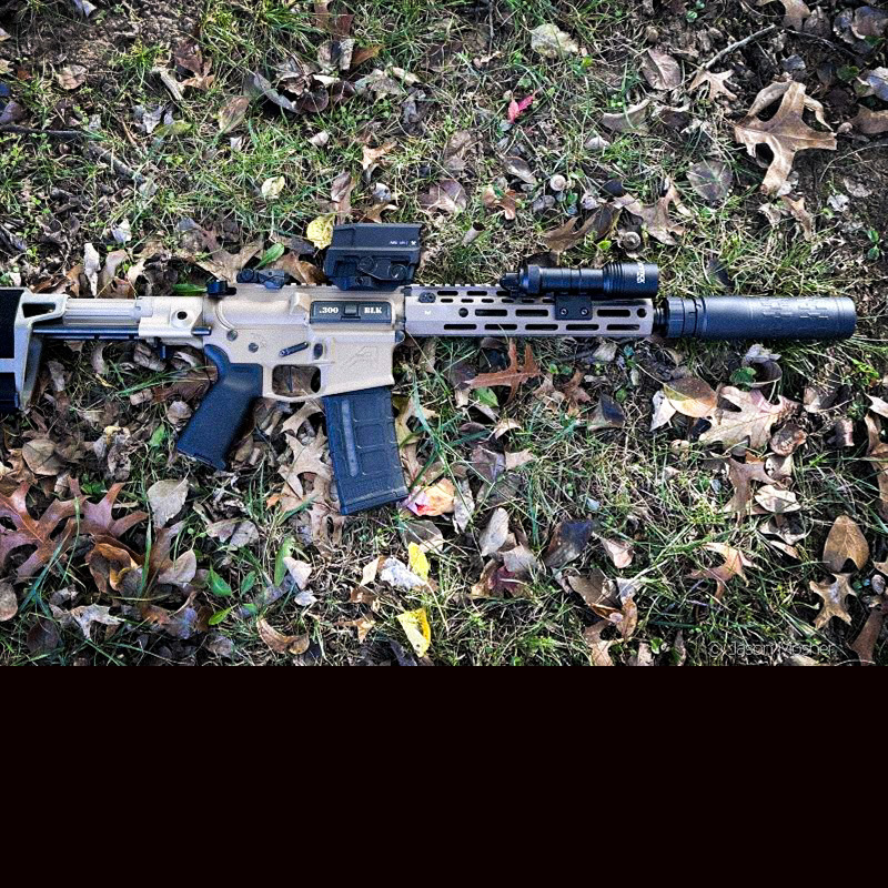 A .tan colored AR-15 rifle laying on the ground with green grass and leaves around it. 