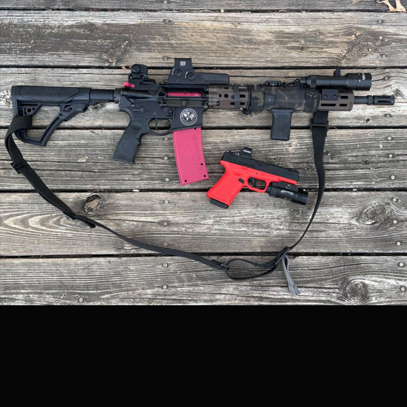 An AR-15 with a training bolt and magazine inserted and a red and black training pistol on a wood floor.