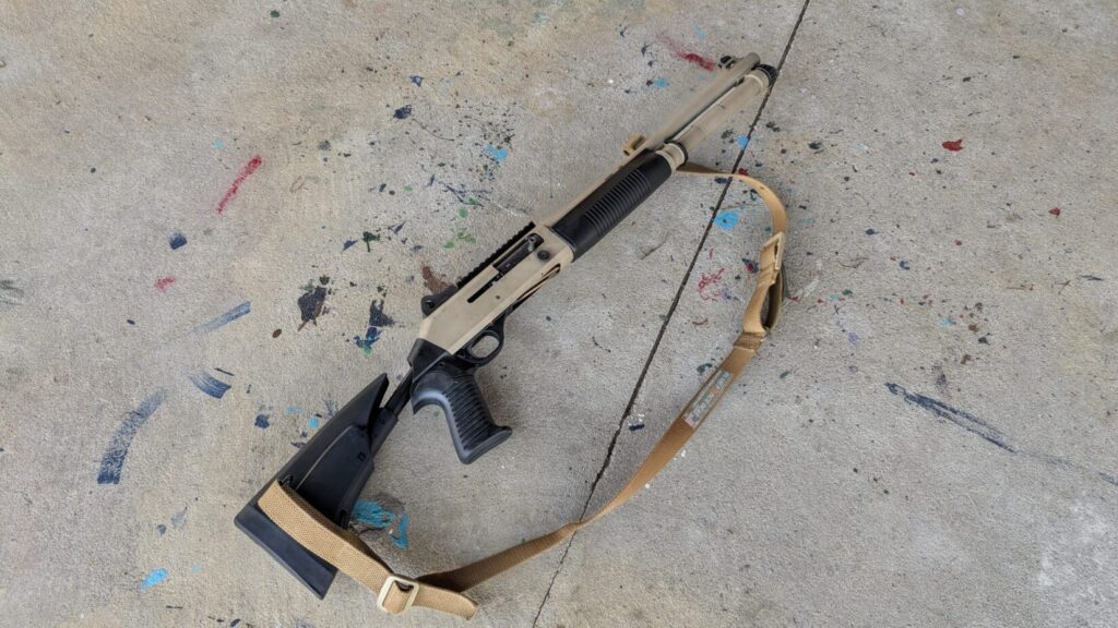 Benelli M4 shotgun in flat dark earth and black, with sling, lying on concrete.