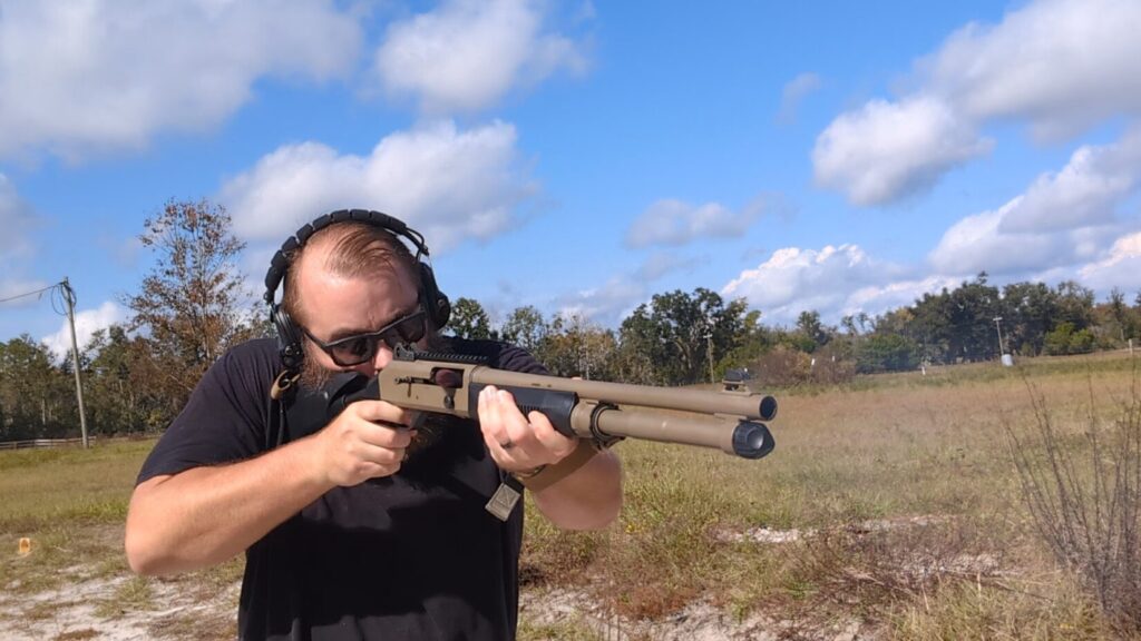 Shooter firing the Benelli M4 shotgun outdoors, with an ejected shell visible.