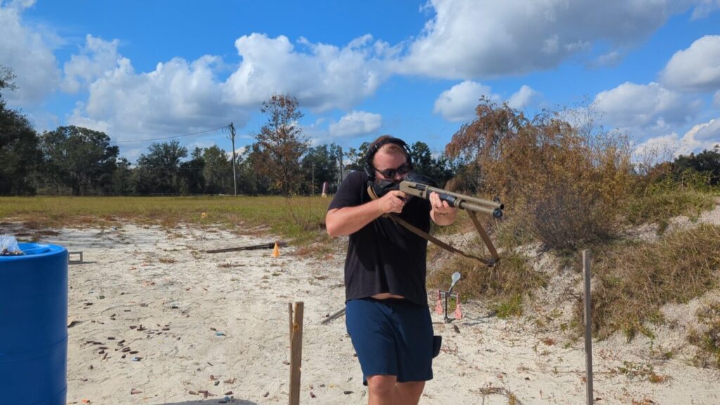 Full-body shot of a shooter engaging targets with the Benelli M4 on a sand-covered range.