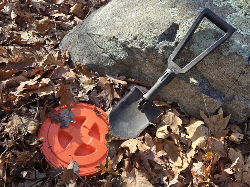 A folding E Tool and the lid of a bucket next to a large rock with leaf debris on the ground