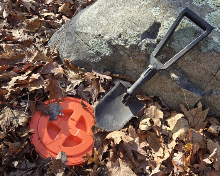 A folding E Tool and the lid of a bucket next to a large rock with leaf debris on the ground