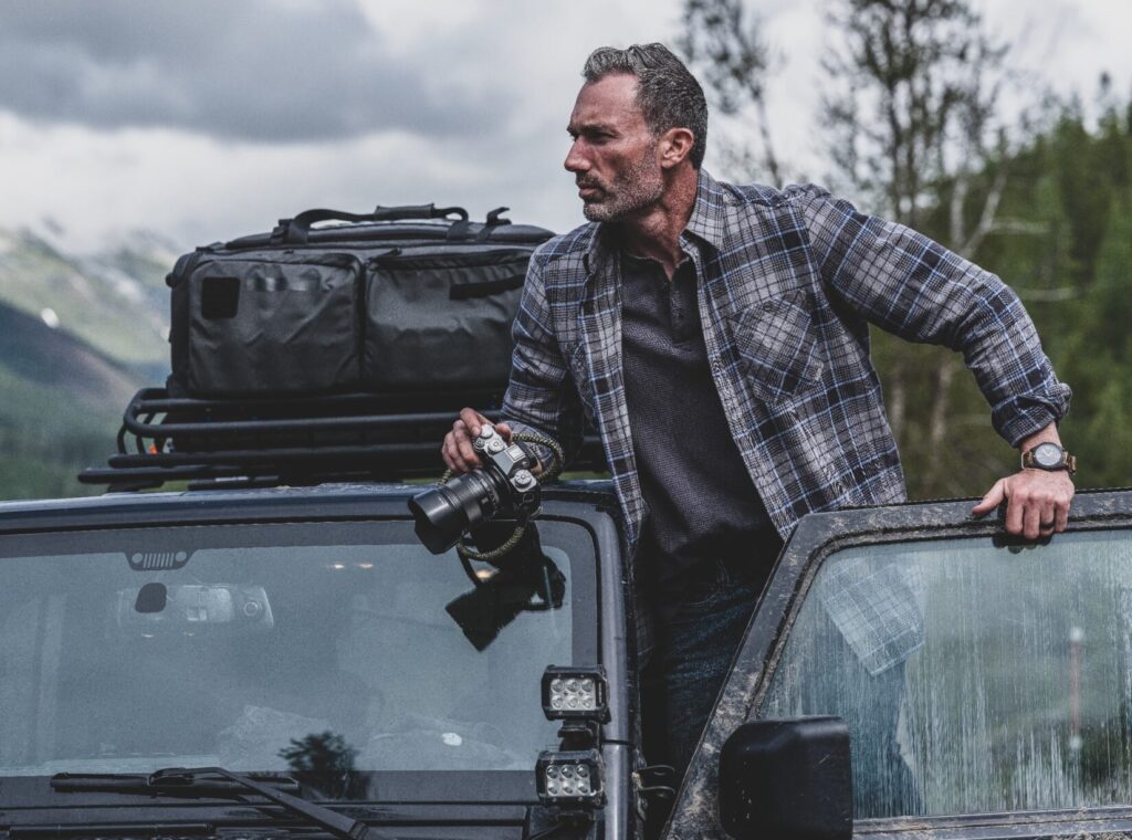 man wearing plaid flannel shirt, climbing out of jeep with camera in hand