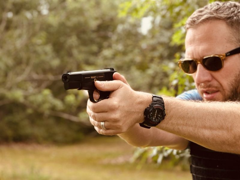 A person shooting the High-Power pistol in a field with trees behind them. 