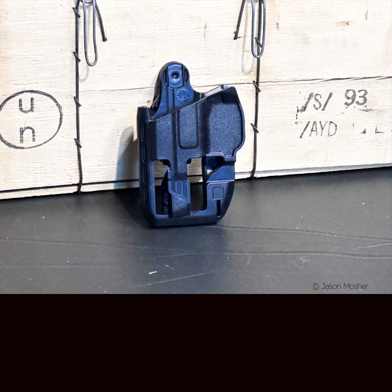 A black holster with areas cutout to reduce weight, leaning up against at wooden box. 