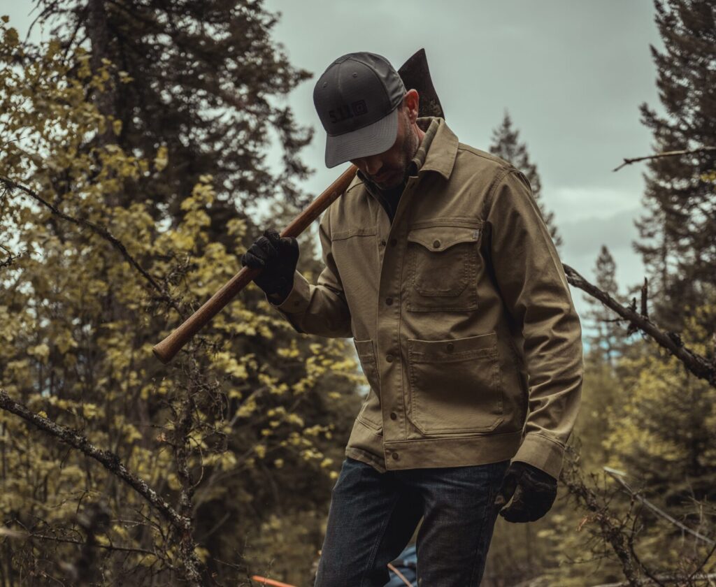 man wearing tan flannel shirt in the woods, carrying an axe