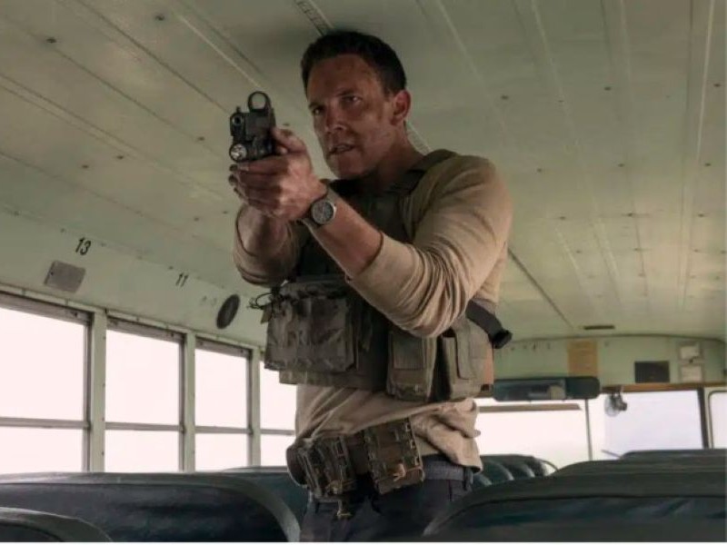A man in tactical clothing, standing on a buss, pointing a pistol at someone.