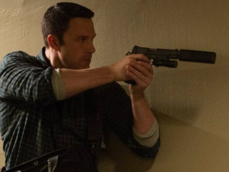 A person standing by a corner, pointing a gun with a silencer on it.