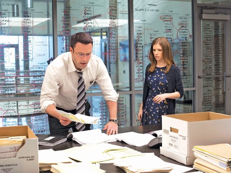 A photo of a movie clip with a man in a white shirt leaning over a table full of books and papers.