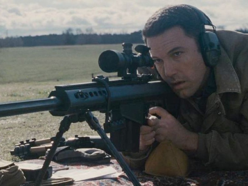 A man in a brown jacket aiming through a rifle scope as he lays on the ground.