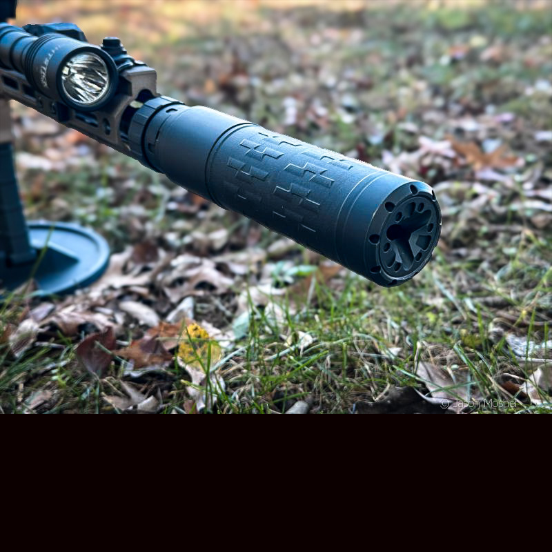 An up close shot of a black silencer attached to the end of a rifle in a field. 