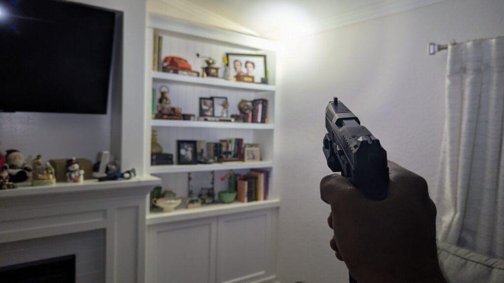 Hand aiming a pistol toward a brightly lit built-in bookshelf in a living room.