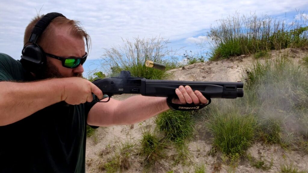 Man with beard and sunglasses shooting a short-barreled shotgun.
